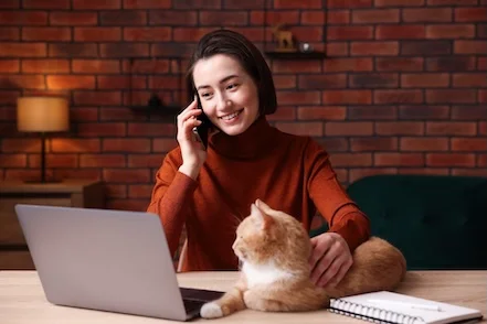 Woman speaking on the phone for customer support with her ginger cat beside her at a laptop